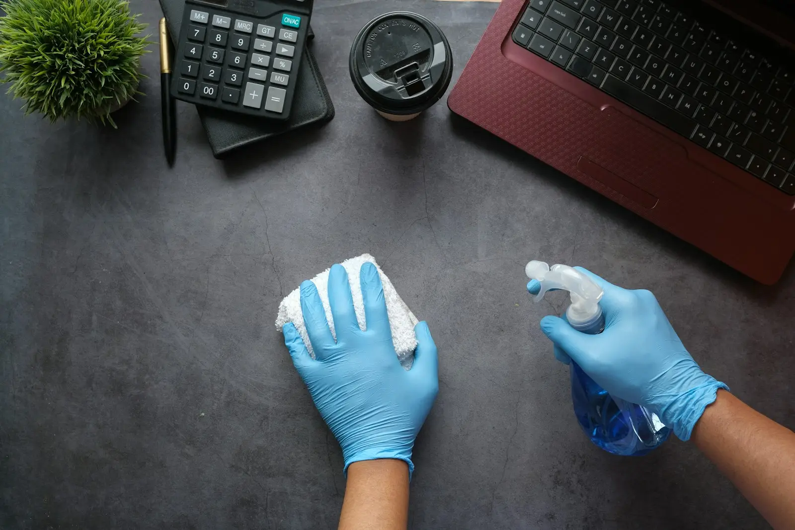 Hände in blauen Handschuhen wischen einen Schreibtisch mit Tuch und Sprühflasche